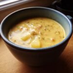 Close-up of a rustic blue bowl filled with creamy, yellow Potato Cheese Soup, featuring visible chunks of potato and herbs.