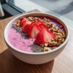 A vibrant pink Strawberry Smoothie Bowl topped with sliced fresh strawberries, chia seeds, and crunchy granola.