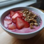 A close-up of a vibrant pink Strawberry Smoothie Bowl topped with fresh sliced strawberries, crunchy granola, and chia seeds.